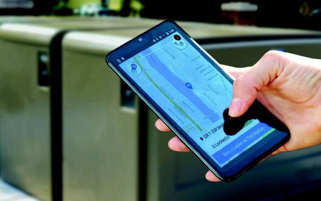 Close-up of a smartphone displaying a digital bike locker app interface, with CycleSafe ProPark® lockers in the background.