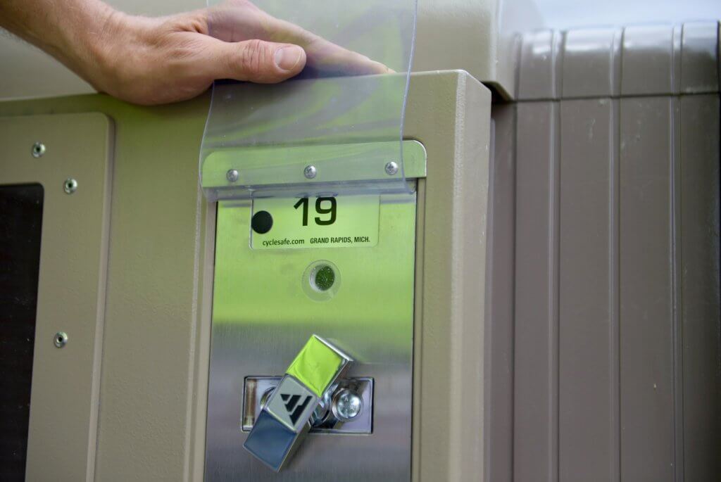 Photograph showing a close-up of a beige CycleSafe ProPark bike locker with a metal t-handle lock and a label marked "19" from Grand Rapids, Michigan. A hand rests on top of the locker, highlighting the lock mechanism and label details.
