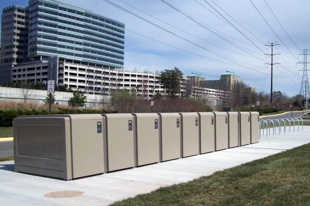 Photograph showing a row of ten beige CycleSafe ProPark® Bike Lockers lined up on a concrete sidewalk next to a grassy area and stainless steel bike racks. Background includes office buildings, trees, and power lines under a partly cloudy sky.