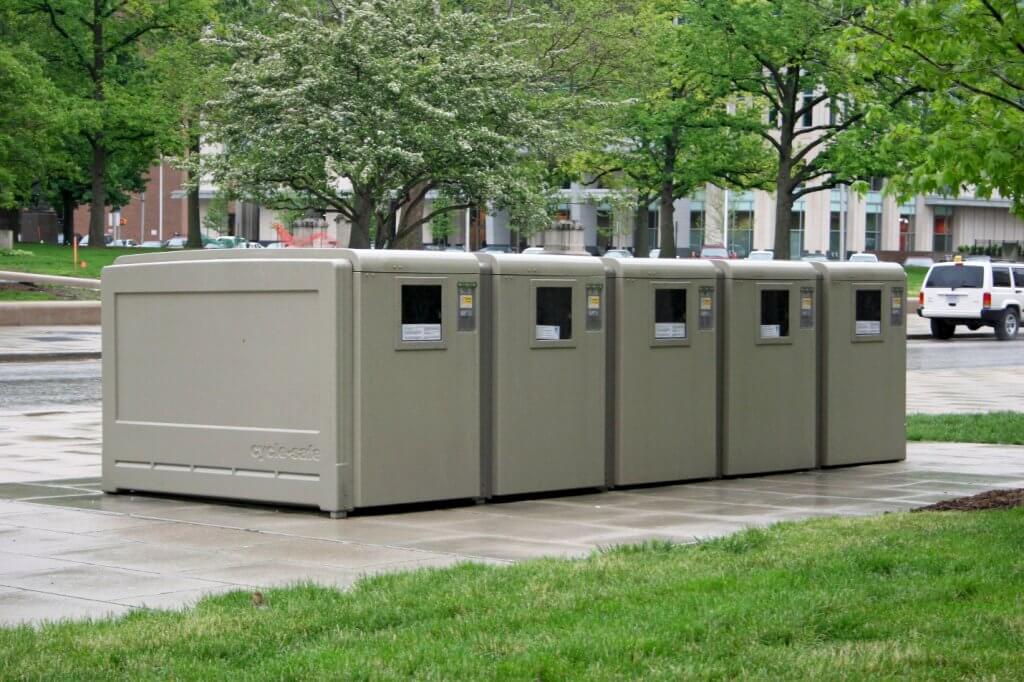 Photograph showing a row of five large, gray ProPark® System Door-View Bike Lockers placed on a paved area near a grassy lawn in an urban park setting. Each locker has a small rectangular window and labels, with trees and buildings visible in the background.