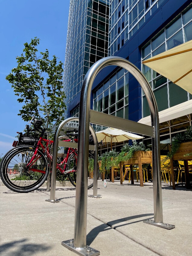 Photograph of a modern outdoor bike rack in front of a commercial building with reflective glass windows and a patio area with tables, chairs, and umbrellas. A red bicycle is parked at the rack, and a tree is visible against a clear blue sky, highlighting an urban setting with amenities for cyclists.