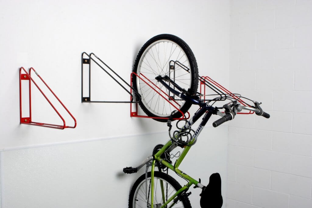 Photograph of a vertical bike storage system mounted on a white wall, featuring red and black metal brackets designed to hold bicycles by their front wheels. One green and black bike is partially lifted into a bracket, demonstrating how the system saves floor space by suspending bikes vertically.