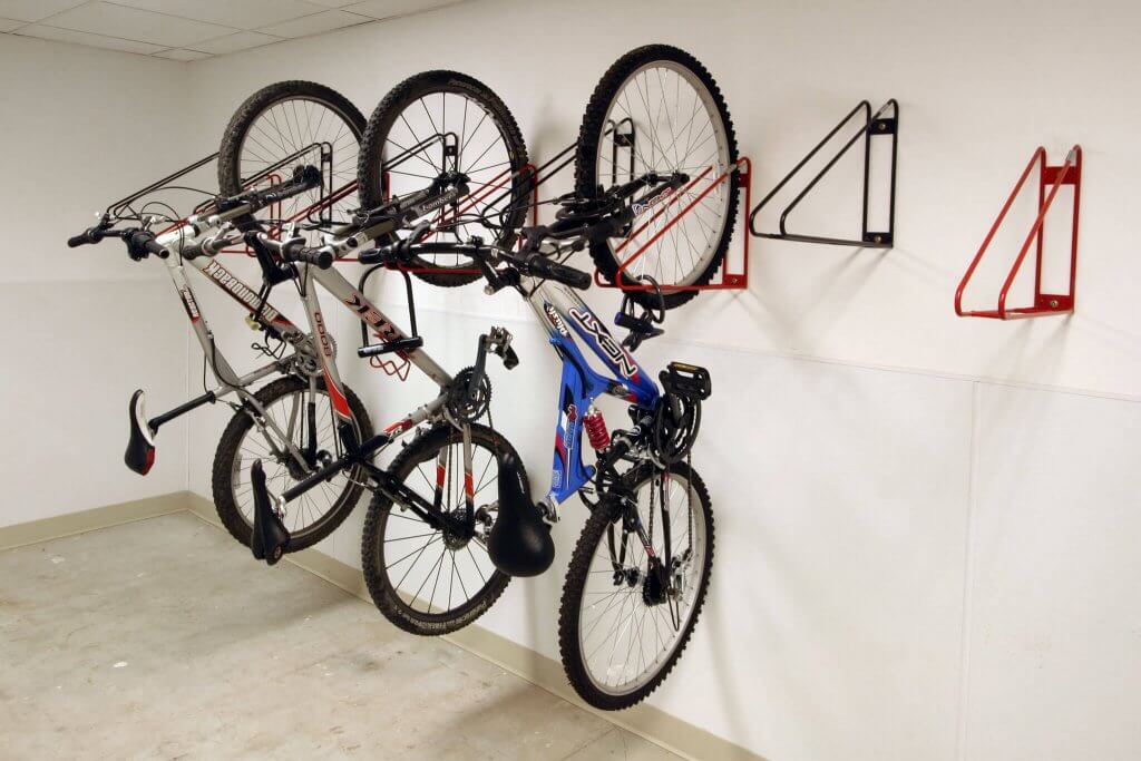 Photograph of three bicycles hanging vertically by their front wheels on red and black CycleSafe wall-mounted bike racks inside a white room with tiled ceiling and concrete floor. The bikes vary in color and design, with two mountain bikes and one road bike, demonstrating an efficient indoor storage solution.