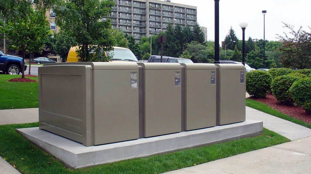 Photograph of four large, beige outdoor ProPark Bike Lockers installed on a concrete platform in a landscaped urban area with grass, bushes, and a high-rise building in the background. Each locker has a small lock and label, indicating secure bike storage.