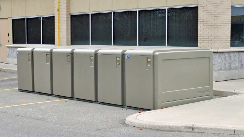 Photograph of a row of six large, gray ProPark® Bike Lockers lined up on a parking lot next to a beige brick building. The lockers are uniform in size and shape, with small labels and locks visible on their front panels, indicating secure bike storage.