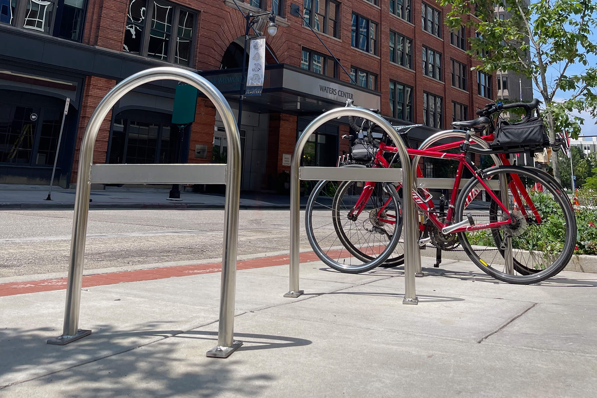 Photograph of CycleSafe bike racks with two empty metal racks and one red bicycle secured to the far rack. Background features a brick building with large windows, a sidewalk, street, and some greenery under a clear sky.
